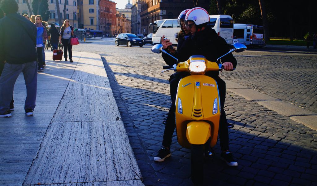 Tourists ride on a yellow Vespa while checking their phones in the streets of Italy.