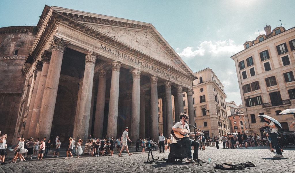 Tourists and locals walk in front of the Pantheon while an artist sings.